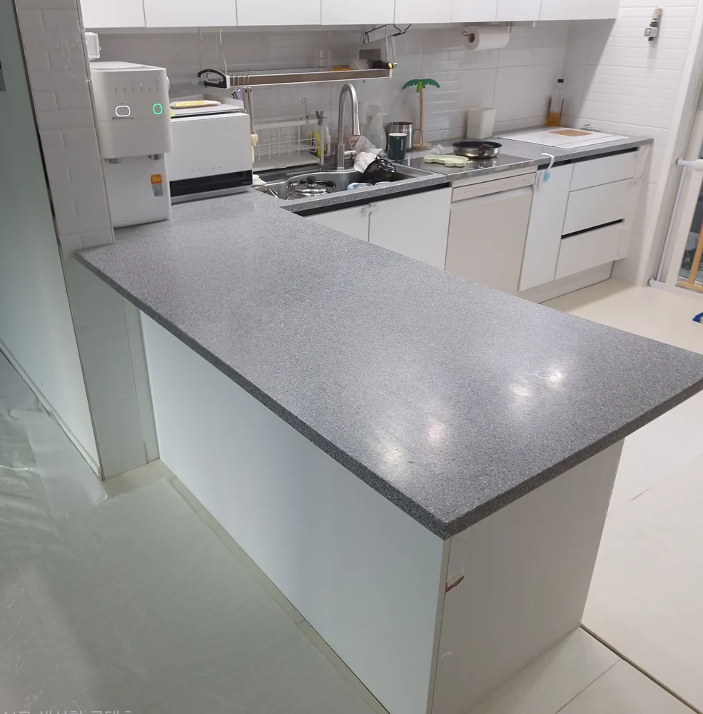 아일랜드식탁 컷팅 시공 현장, 바닥에 보양 비닐을 깐 주방에서 흰색 하부장 위로 길게 나온 회색 알갱이 ㄴ자 상판